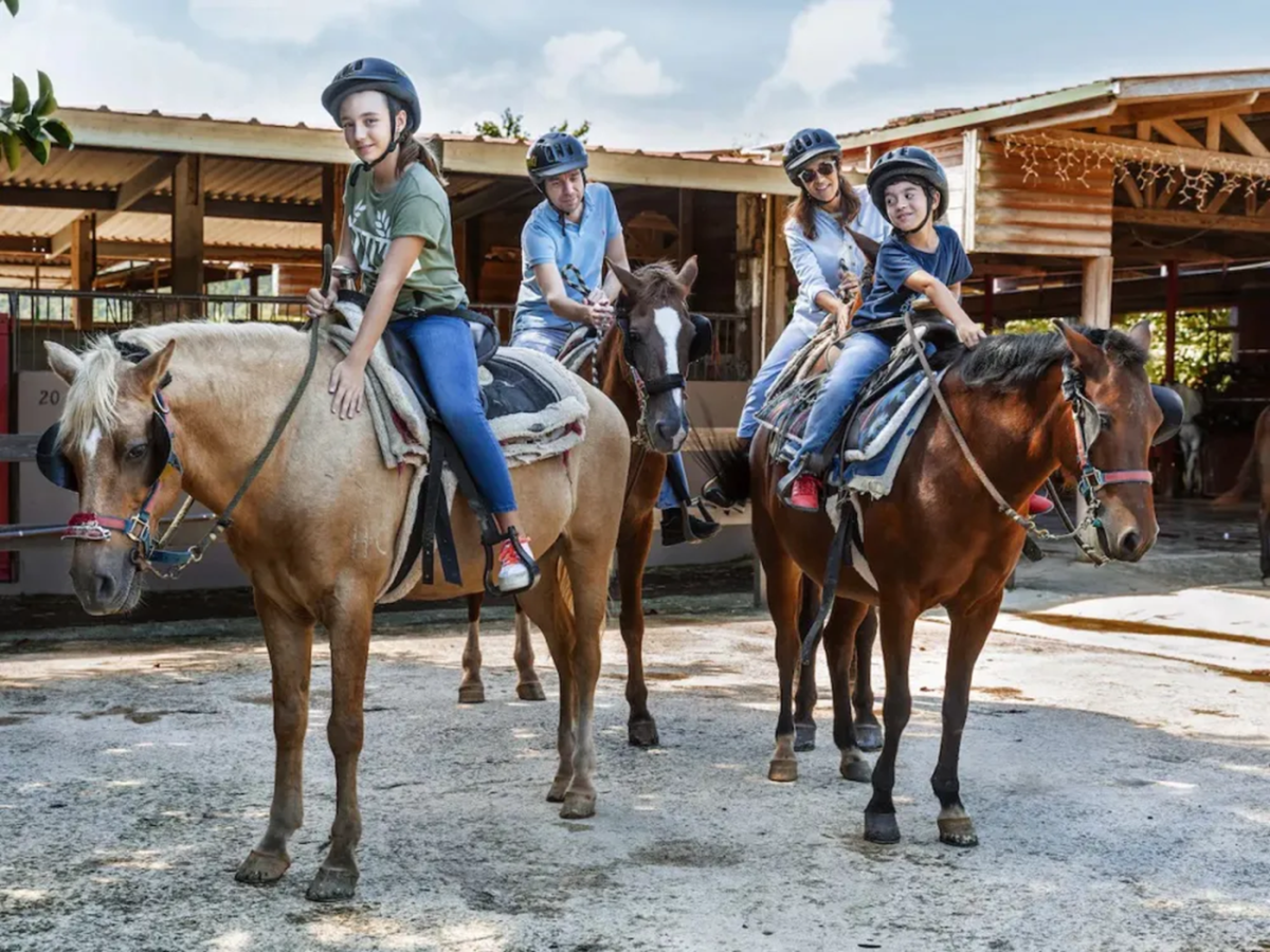 Three people riding horses at a stable, wearing helmets and casual attire.