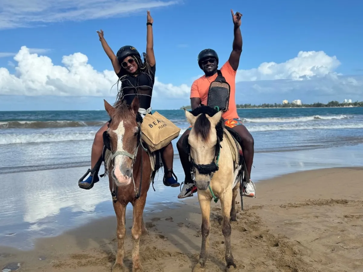 Two people on horses at the beach raising arms with ocean and blue sky in the background.