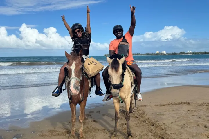Two people on horses at the beach raising arms with ocean and blue sky in the background.