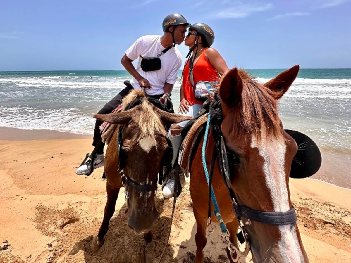 Couple wearing helmets kissing on horseback at the beach with waves in the background.