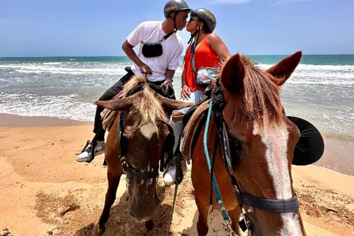 Couple wearing helmets kissing on horseback at the beach with waves in the background.