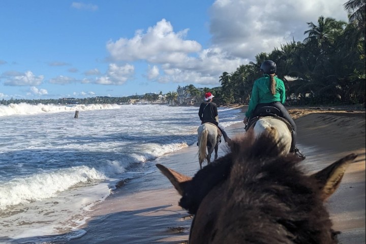 Two people horseback riding on a beach near the ocean under a blue sky with clouds.