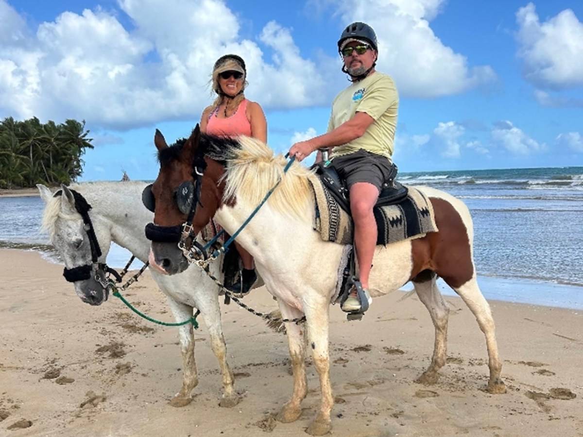 Two people riding horses on a beach with blue sky and palm trees in the background.