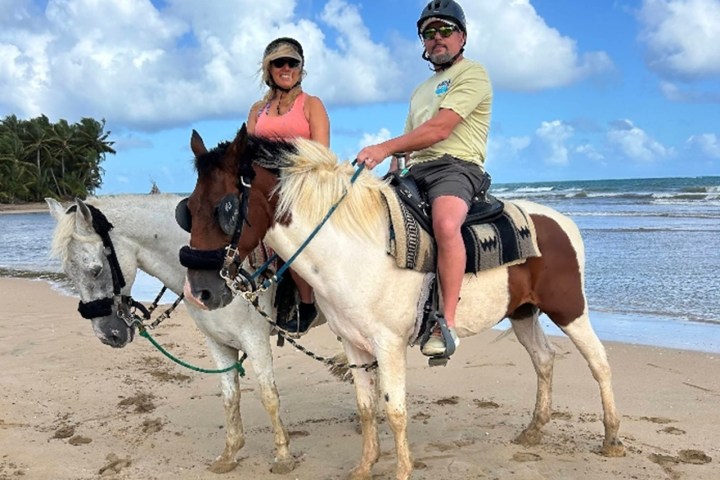 Two people riding horses on a beach with blue sky and palm trees in the background.