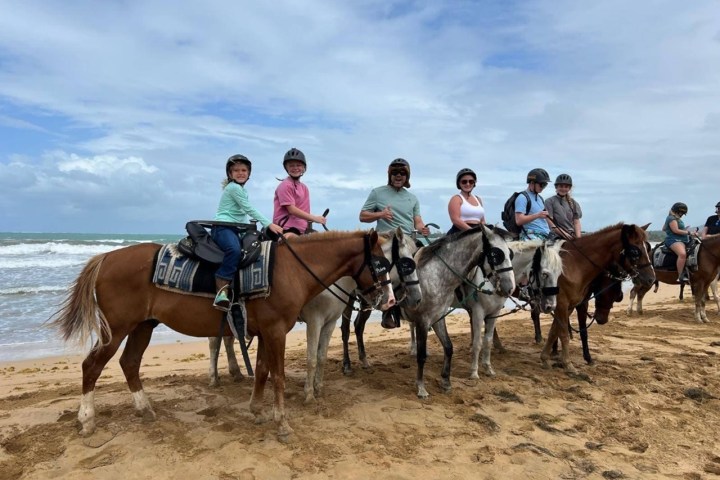 Group of people on horseback at the beach, wearing helmets.