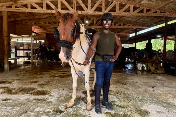 Person in helmet and glasses stands smiling with a horse in a wooden stable.