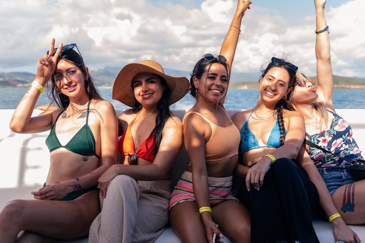 Five women in swimwear smiling and posing on a boat in sunny weather.