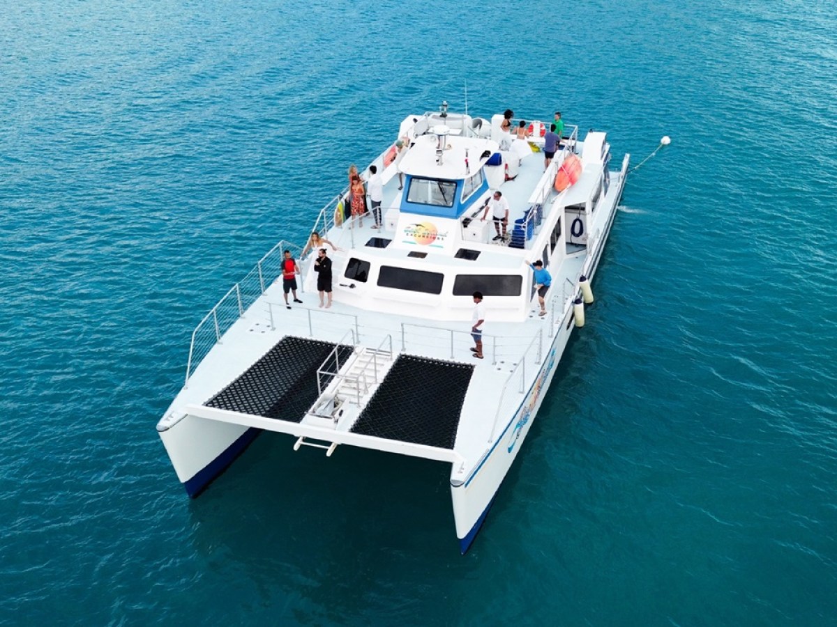 Catamaran with people on deck, sailing on calm blue water.