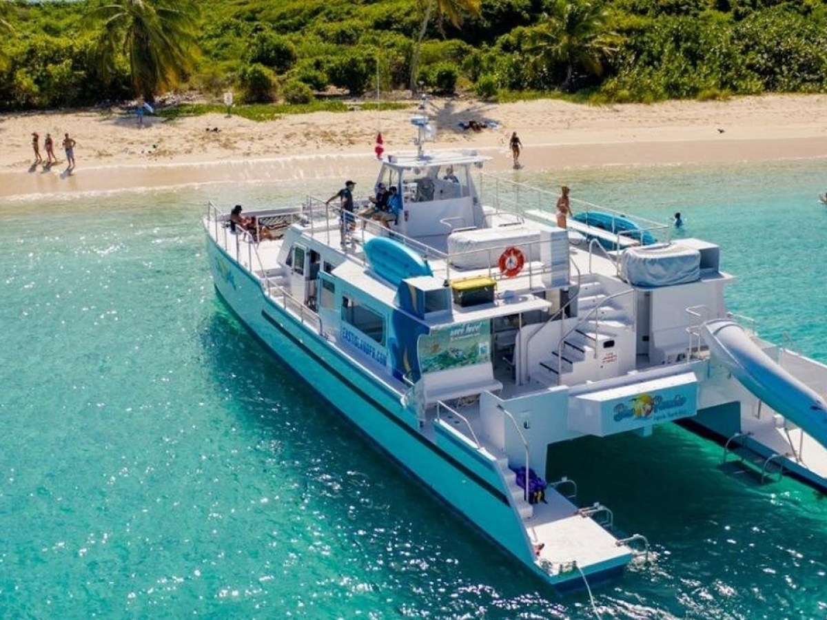 People near a large boat with a slide, anchored by a tropical beach.