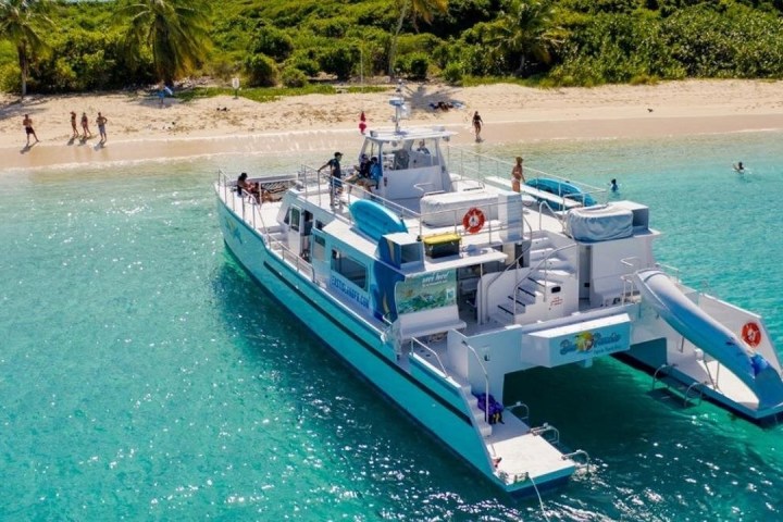 People near a large boat with a slide, anchored by a tropical beach.