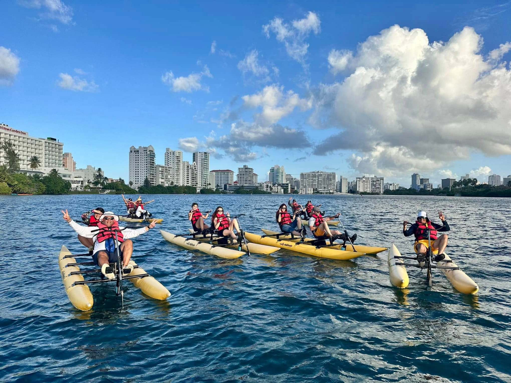Group kayaking on pedal boats in a city harbor, wearing red life vests under a clear blue sky.