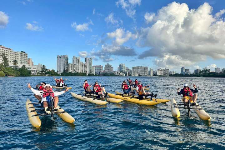 Group kayaking on pedal boats in a city harbor, wearing red life vests under a clear blue sky.