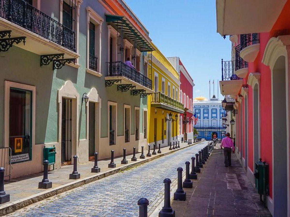 Colorful street with pastel buildings and balconies, leading to a distant blue building under a clear sky.