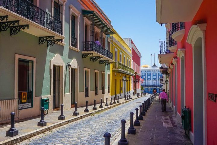 Colorful street with pastel buildings and balconies, leading to a distant blue building under a clear sky.