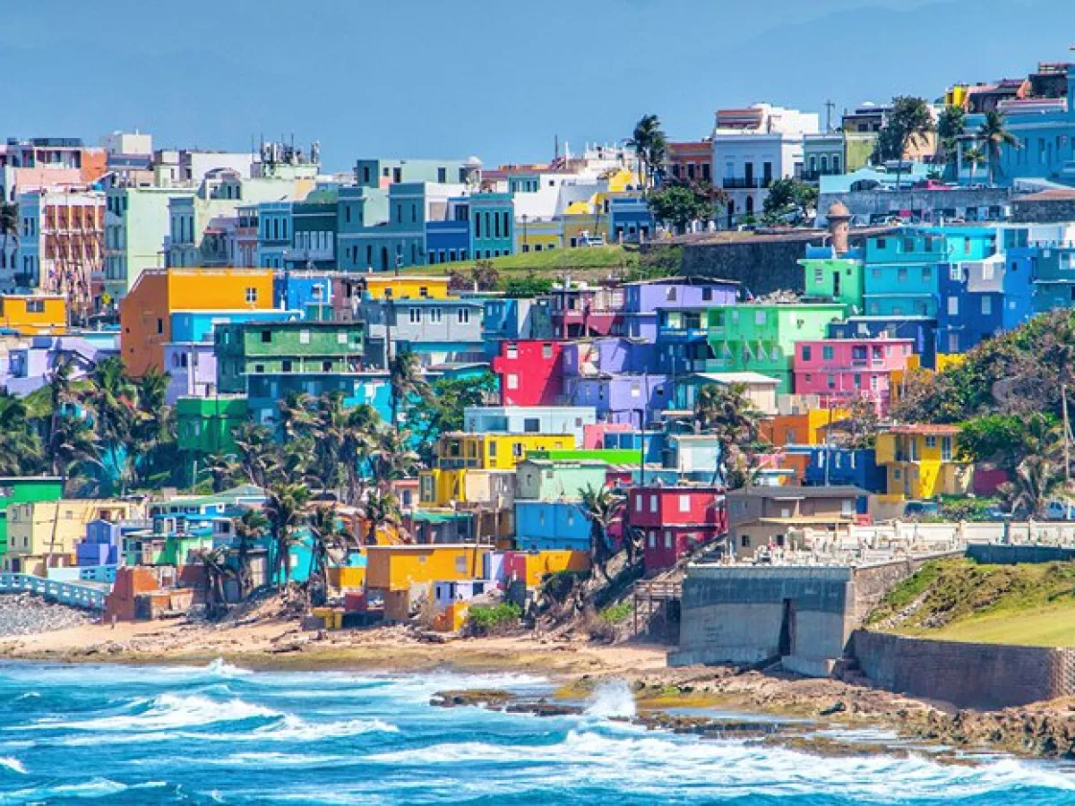 Colorful hillside buildings near a coast with waves crashing onto rocks in the foreground.