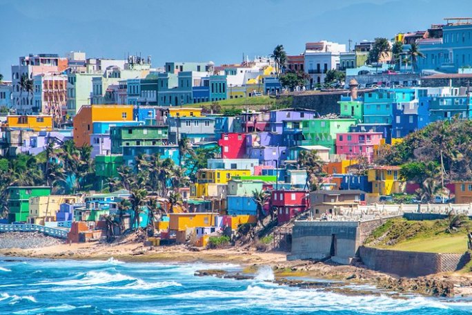 Colorful hillside buildings near a coast with waves crashing onto rocks in the foreground.