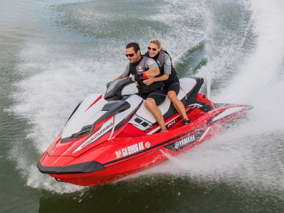 Two people riding a red Yamaha jet ski on water, creating a spray trail.