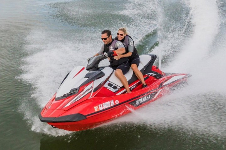 Two people riding a red Yamaha jet ski on water, creating a spray trail.