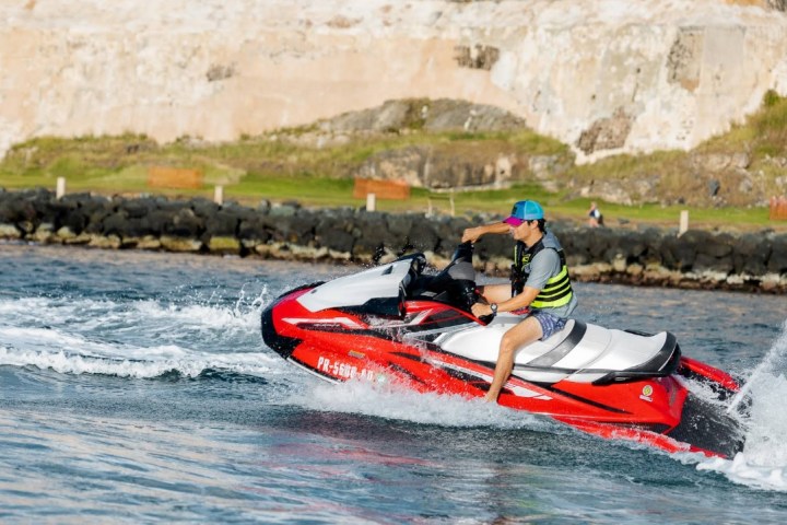 Man in life vest riding red jet ski on water near rocky shoreline.