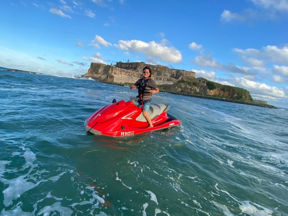 Person riding a red jet ski on the ocean with a historic fort in the background under a blue sky with clouds.