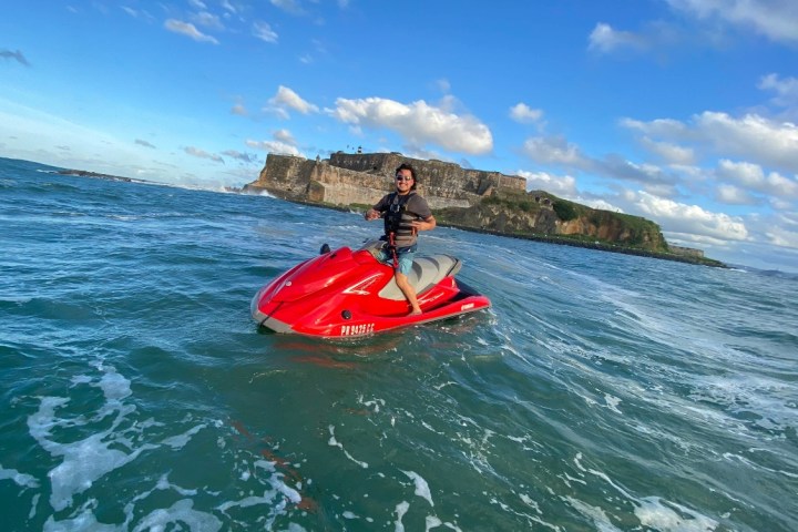 Person riding a red jet ski on the ocean with a historic fort in the background under a blue sky with clouds.
