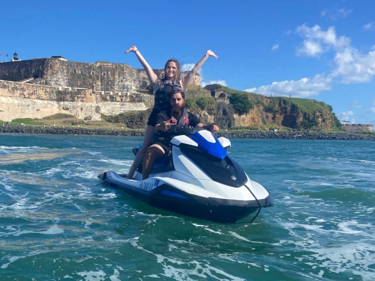 Two people on a jet ski in front of a historic stone fort, with blue sky and ocean waves.