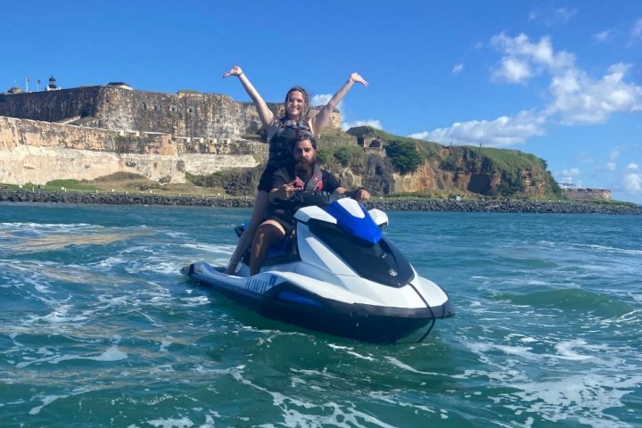 Two people on a jet ski in front of a historic stone fort, with blue sky and ocean waves.
