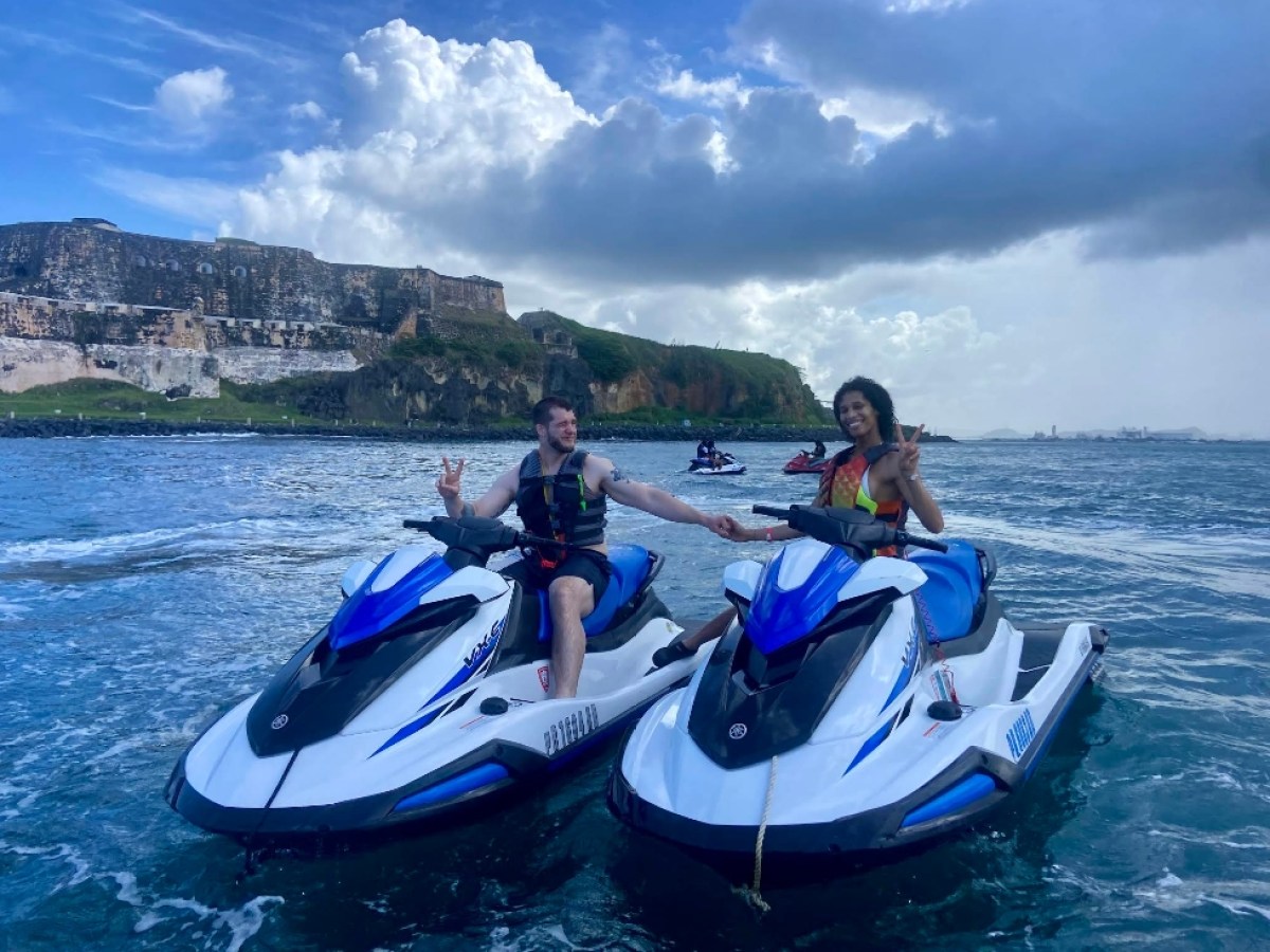 Two people on jet skis holding hands near a historic fortress on a partly cloudy day.