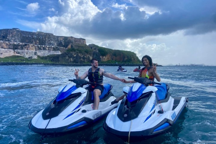 Two people on jet skis holding hands near a historic fortress on a partly cloudy day.