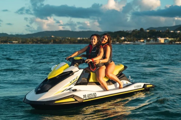 Two people on a jet ski in the ocean, with a distant coastline and cloudy sky in the background.