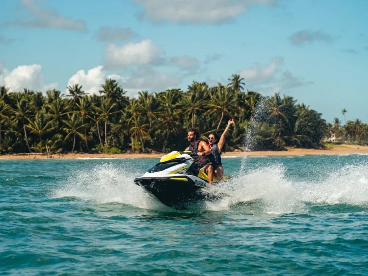 Two people riding a jet ski near a tropical beach with palm trees in the background.