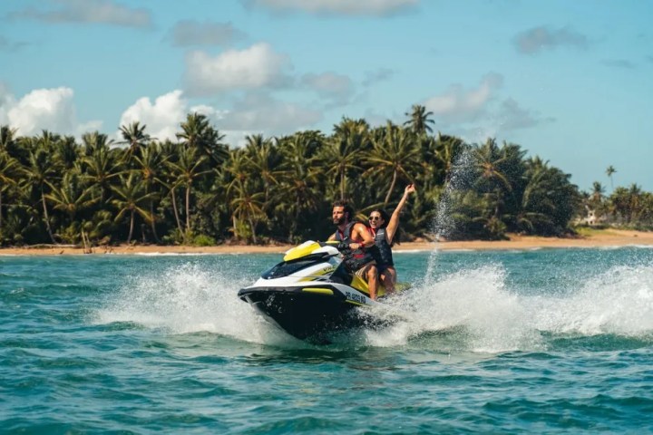 Two people riding a jet ski near a tropical beach with palm trees in the background.