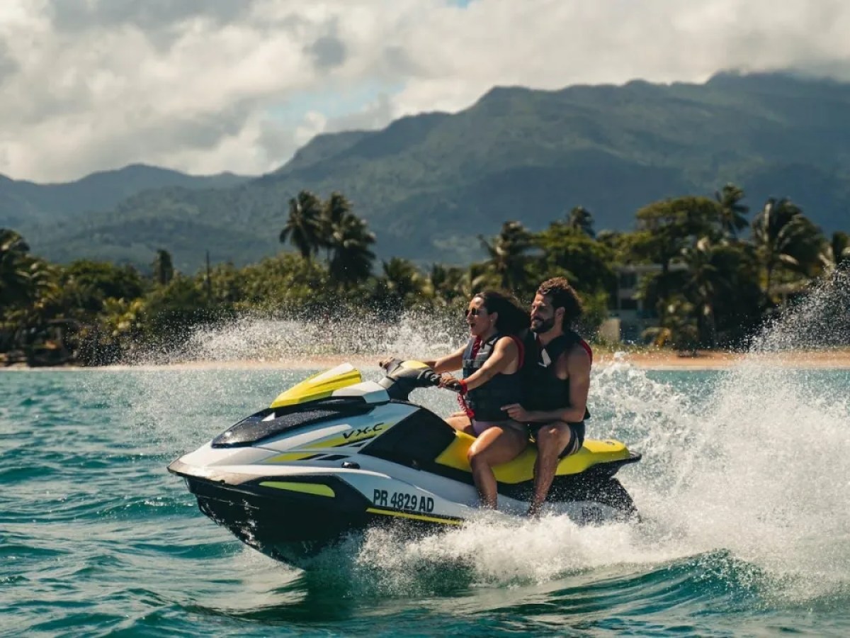 Two people riding a yellow jet ski on the ocean with mountains in the background.
