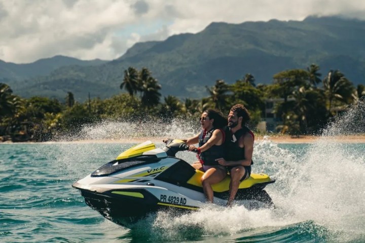 Two people riding a yellow jet ski on the ocean with mountains in the background.