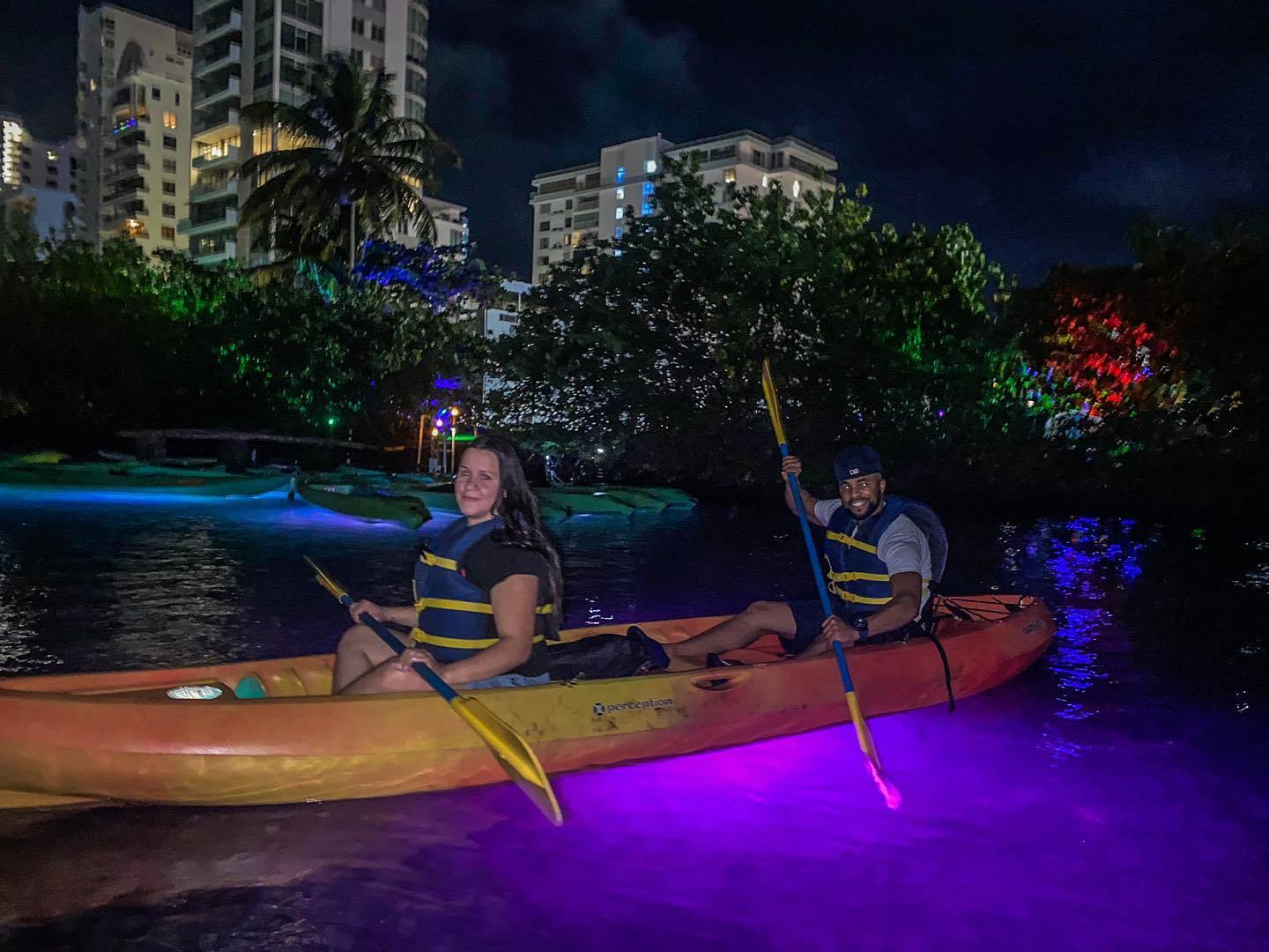 Two people kayaking at night with glowing paddles and water, buildings and trees in the background.