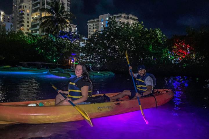 Two people kayaking at night with glowing paddles and water, buildings and trees in the background.