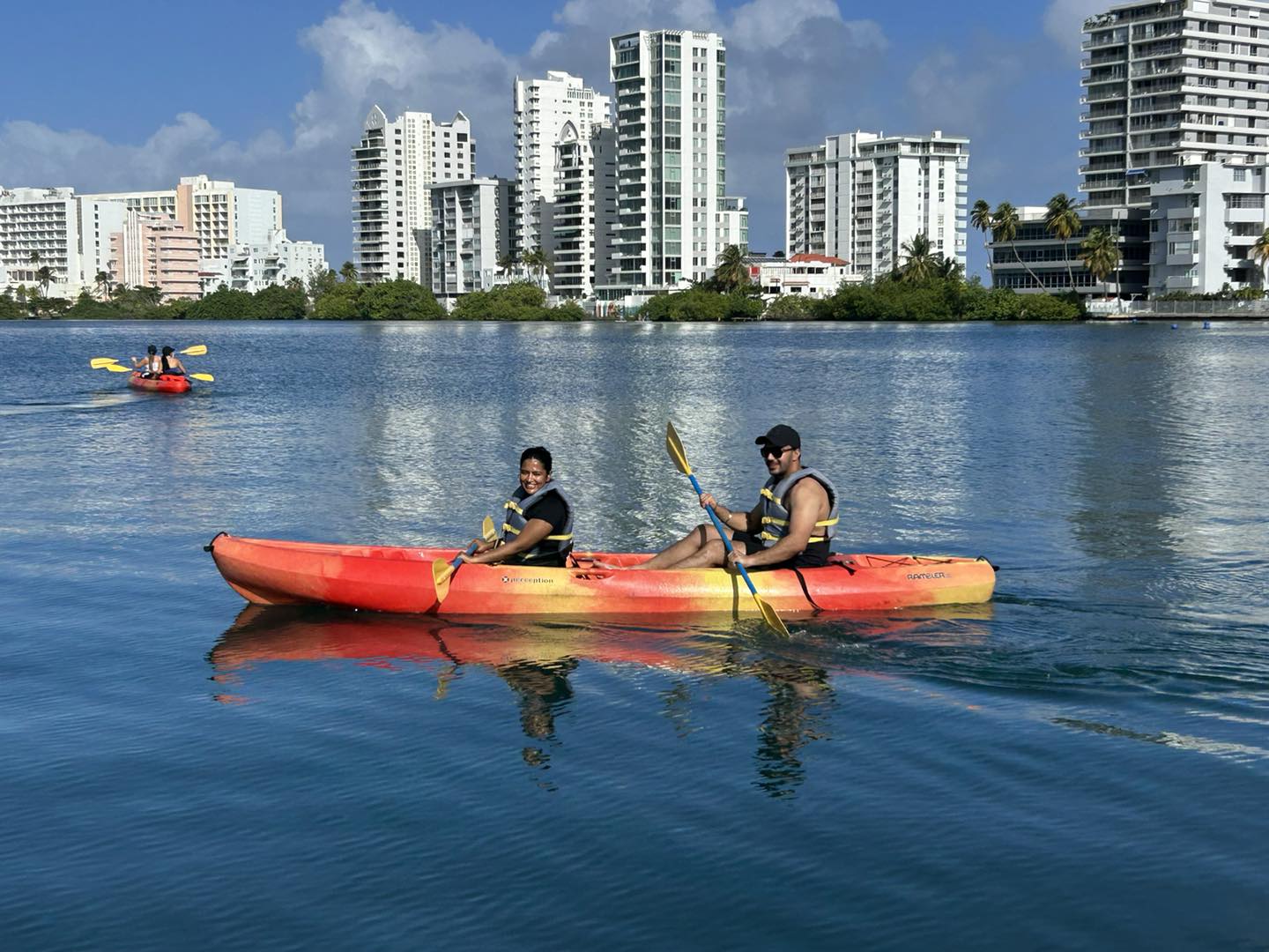 Two people kayaking on calm water with city skyline in the background.