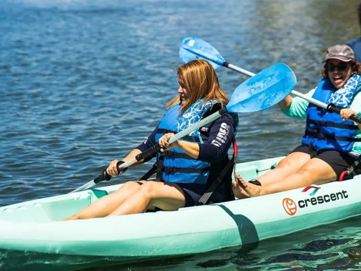 Two smiling women kayaking in blue life vests, with one person standing behind them.