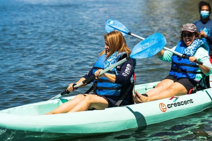 Two smiling women kayaking in blue life vests, with one person standing behind them.