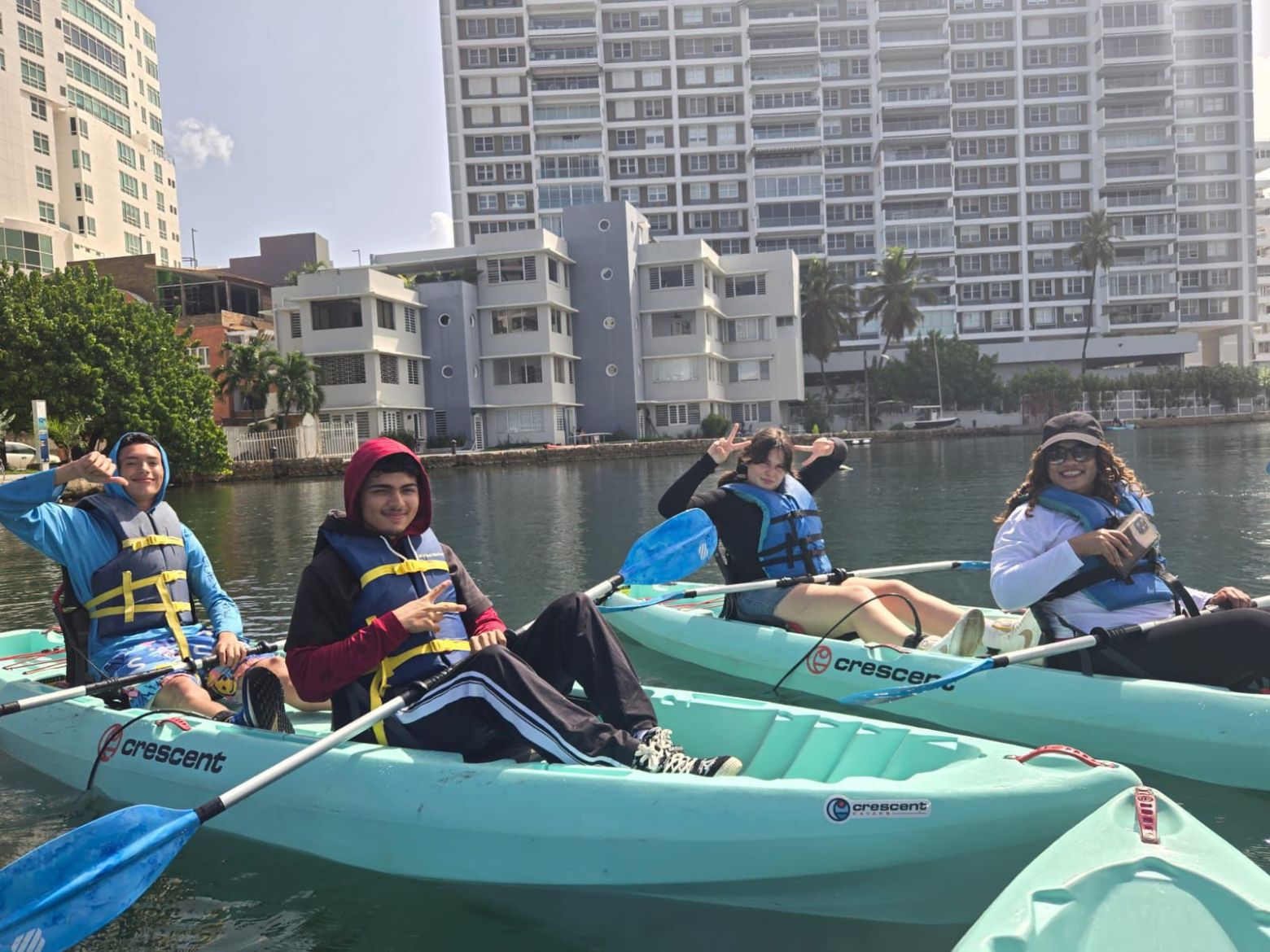 Four people kayaking on a calm urban waterway, with tall buildings in the background.
