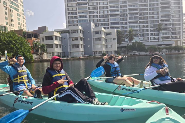 Four people kayaking on a calm urban waterway, with tall buildings in the background.