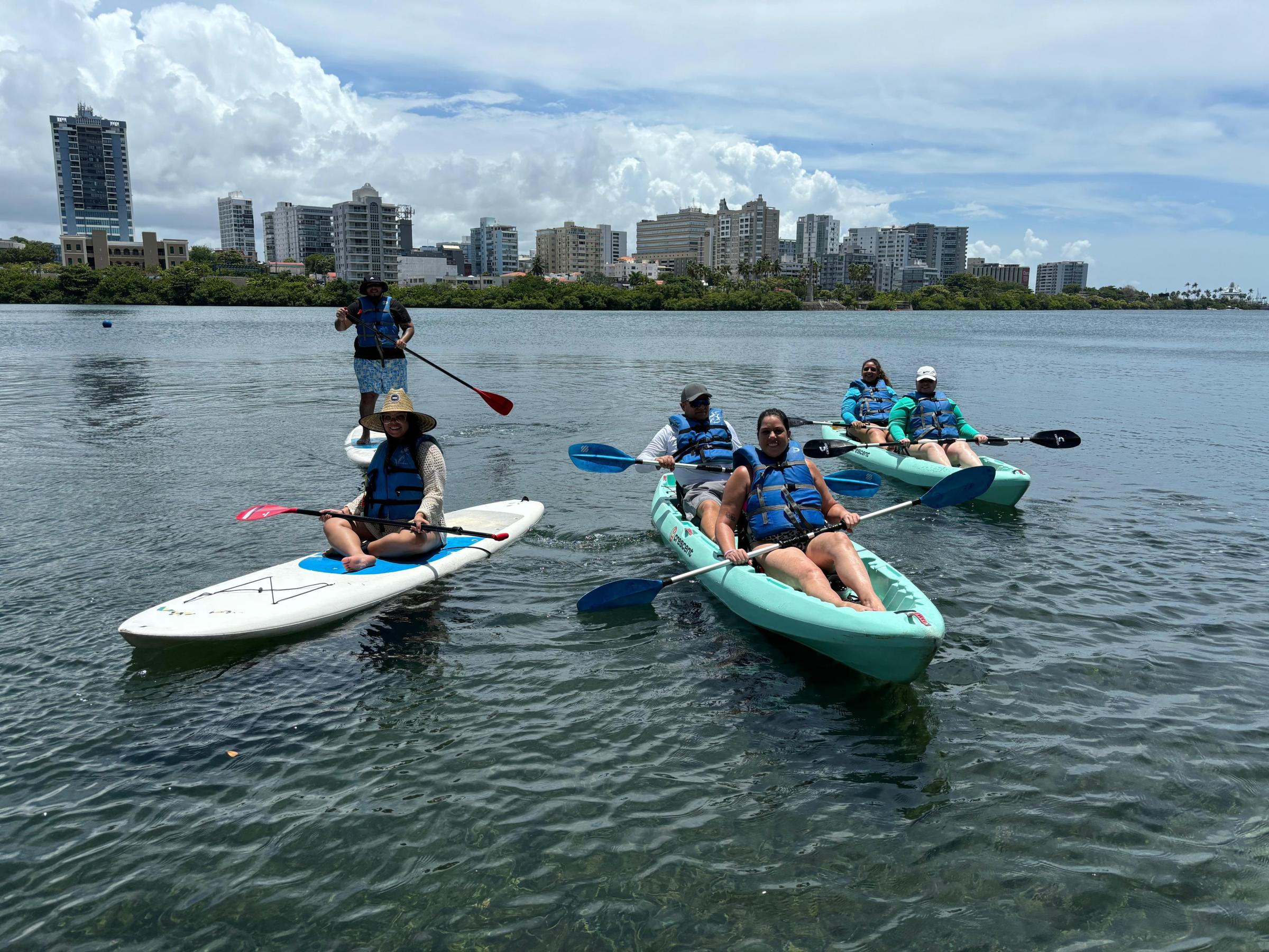 People kayaking and paddleboarding in a calm water body with city skyline in the background.