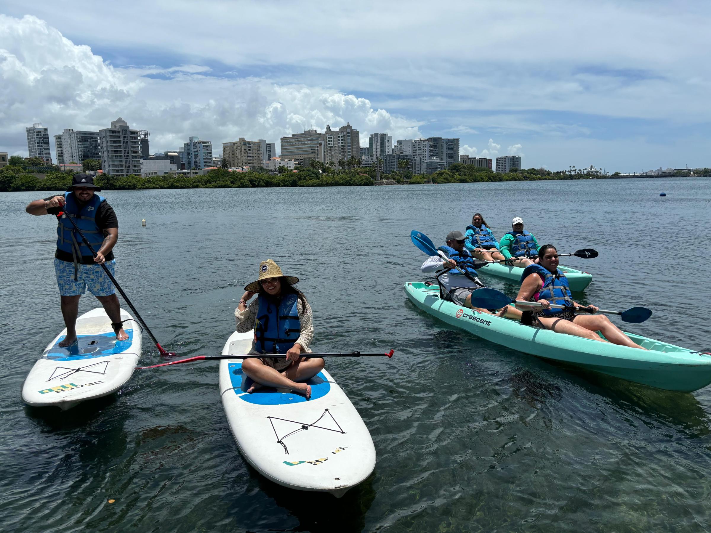 People kayaking and paddleboarding on a sunny day with city skyline in the background.