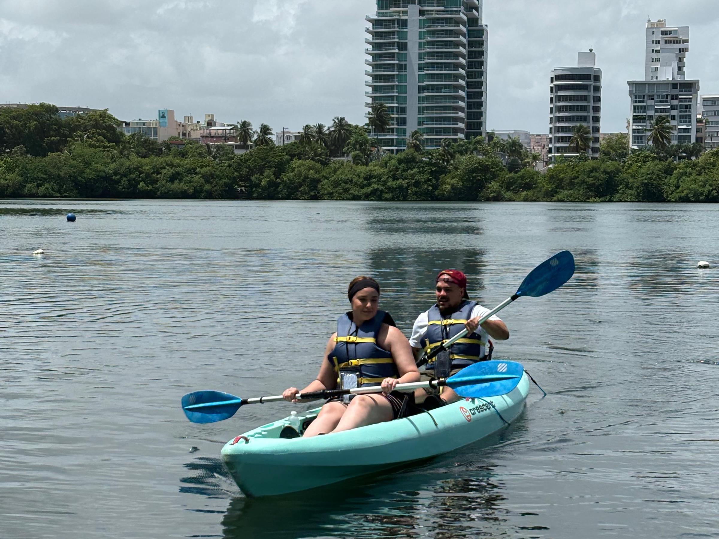 Two people kayaking on calm water with buildings in the background.