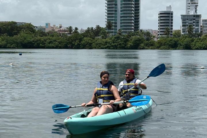 Two people kayaking on calm water with buildings in the background.