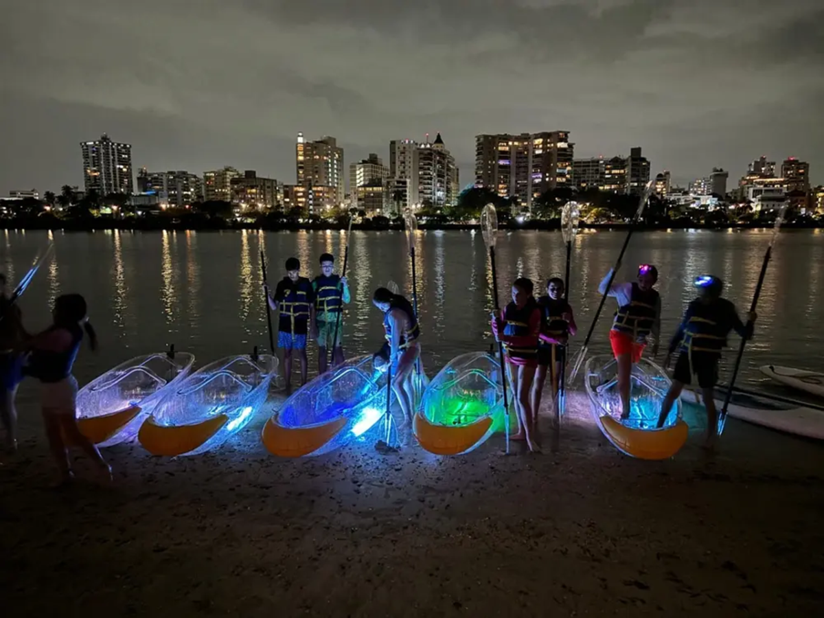 Group with lit kayaks on beach at night, city skyline in background.