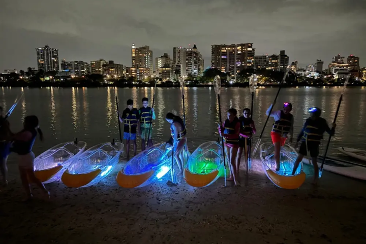 Group with lit kayaks on beach at night, city skyline in background.