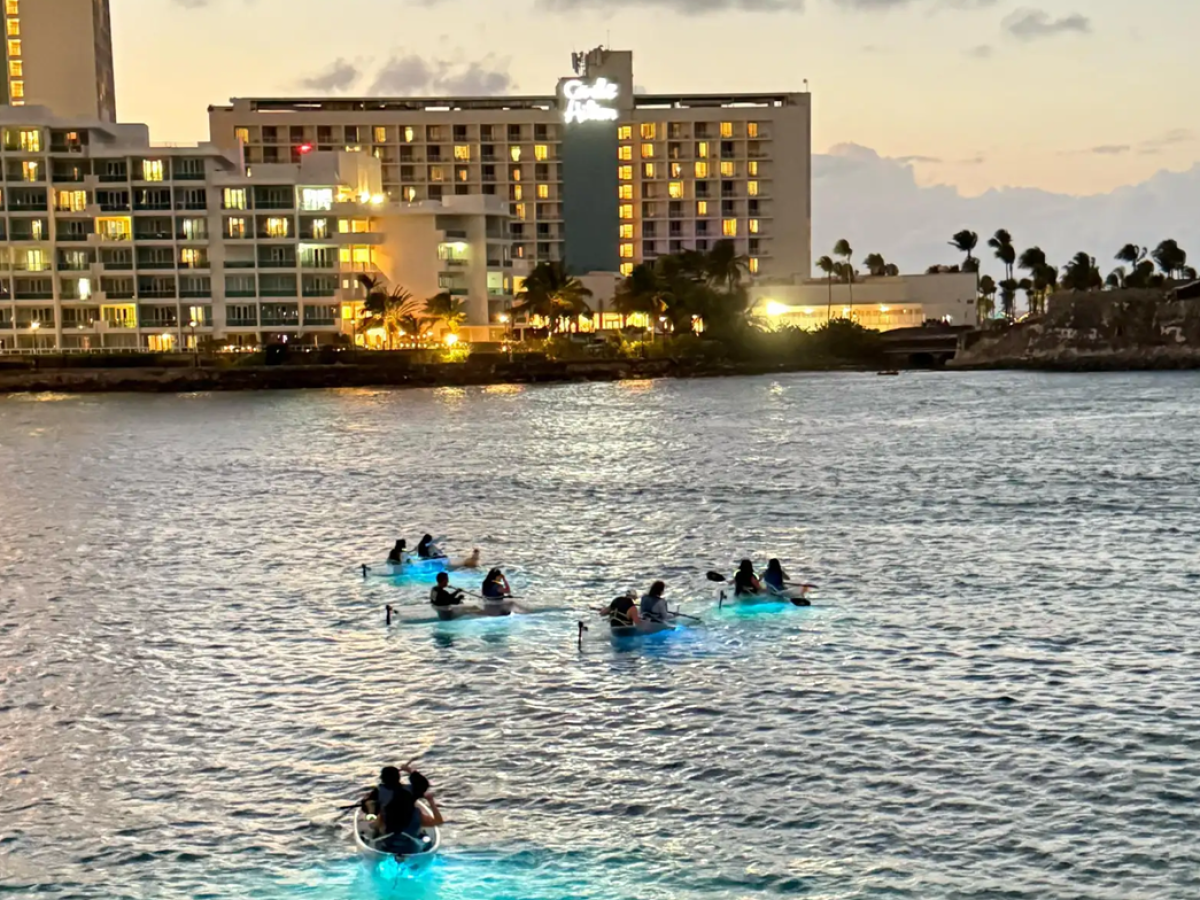 People kayaking on water with illuminated hulls near lit hotels at dusk.