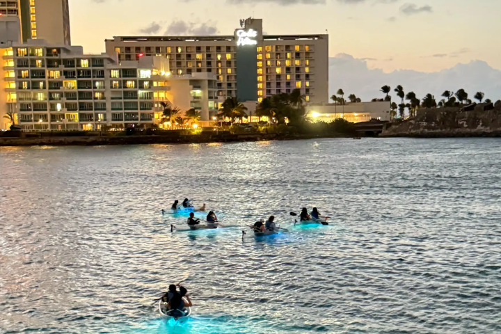 People kayaking on water with illuminated hulls near lit hotels at dusk.