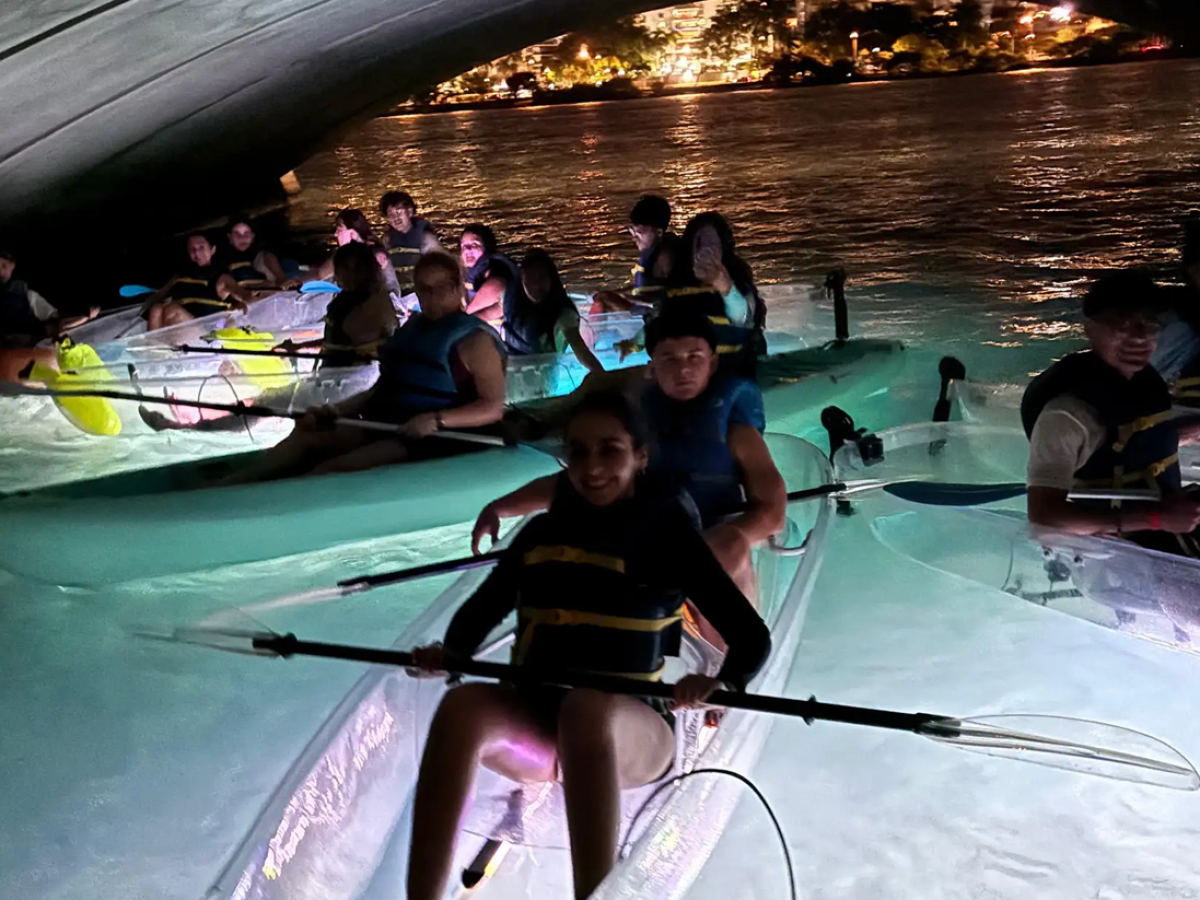 Group kayaking at night under a bridge with illuminated water.
